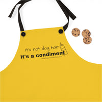 It's a Condiment Mustard Apron - Image description - an adult wearing the mustard colored yellow apron with saying It's not dog hair it's a condiment with an outline of a condiment  bottle with 3 chocolate chip cookies. The black neck time straps are attached with black buttons.